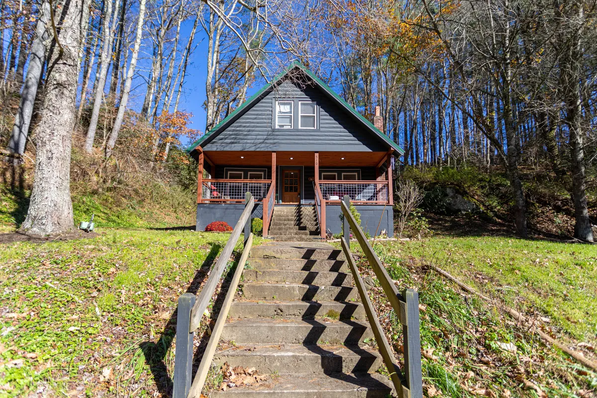 This image shows the front view of the cabin with a staircase leading to the entrance, a gabled roof, and the cabin nestled among lush trees. The porch offers a welcoming spot to relax and take in the sights and sounds of the surrounding nature, reinforcing the connection between the cozy retreat and its picturesque setting.