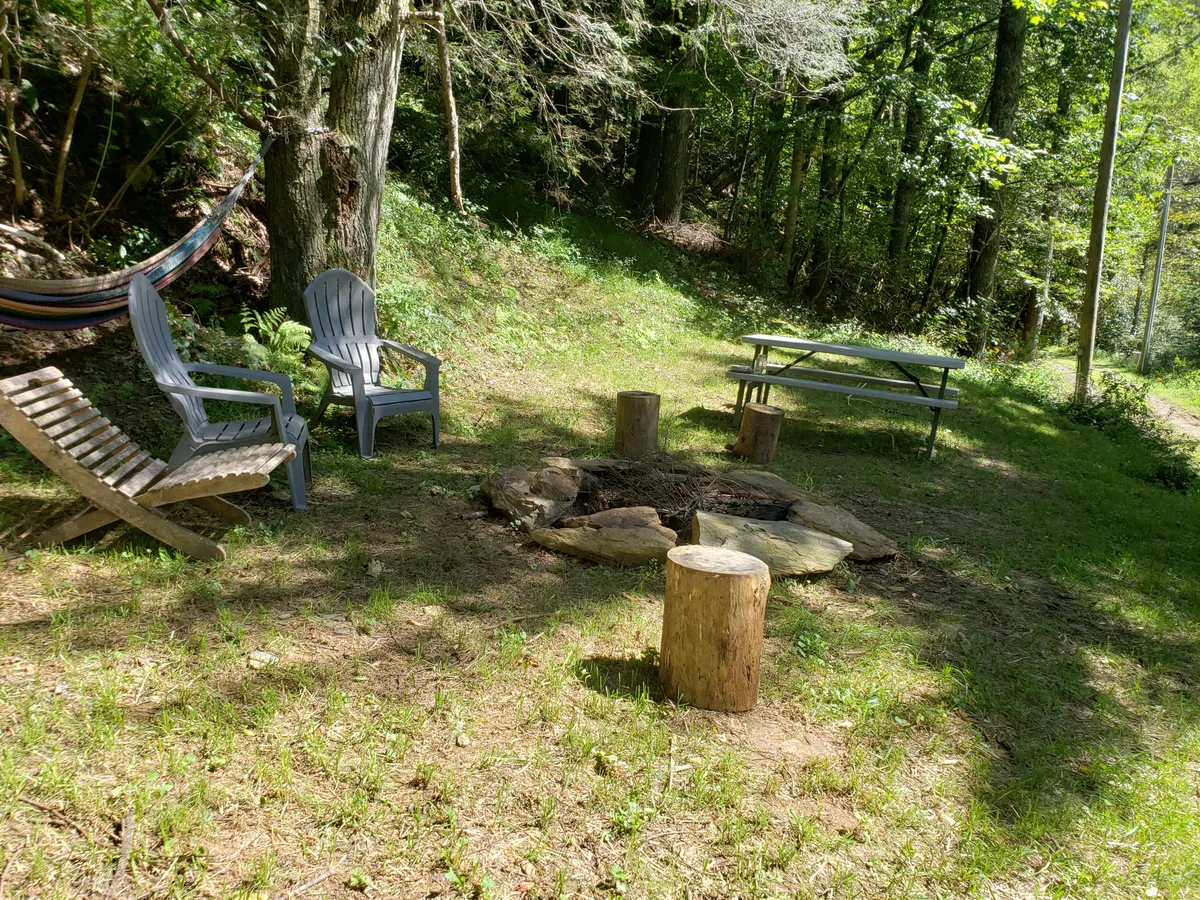 An inviting outdoor scene featuring a fire pit encircled by Adirondack chairs, a rustic picnic table, and a hammock strung nearby. The area is shaded by trees and radiates a communal, cozy vibe, perfect for stargazing or sharing meals after a day out on the trails.