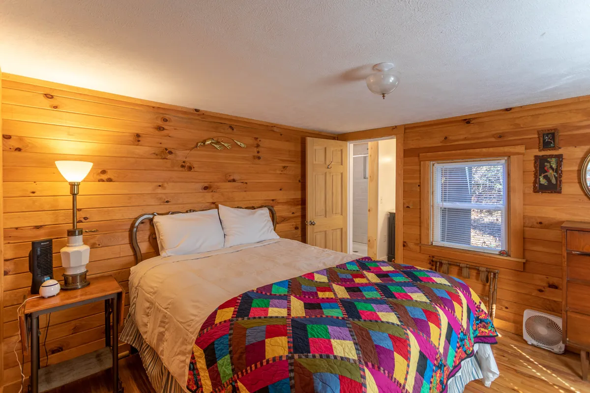 A bedroom featuring a bed with a colorful quilt, warm wooden walls, a glowing lamp, and a window letting in soft natural light. The room radiates warmth and intimacy, making it an inviting sanctuary under the cabin’s charming eaves.