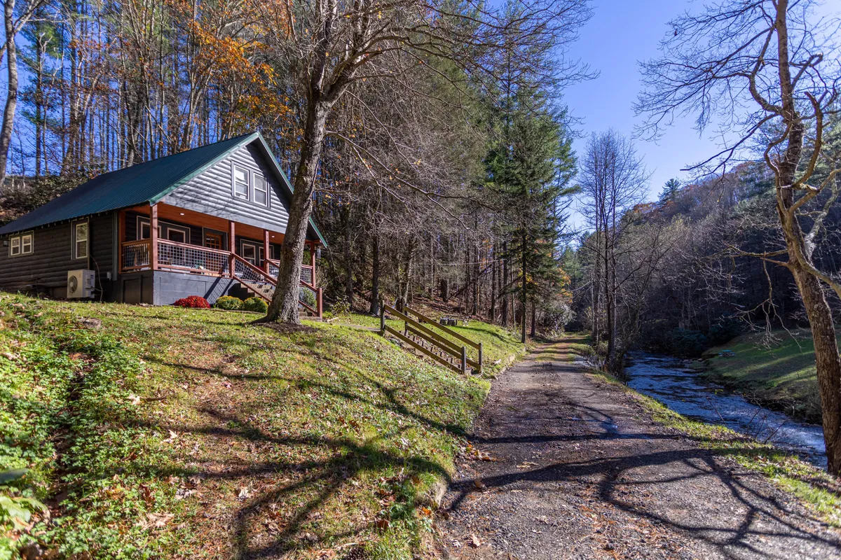 A scenic exterior view of the cabin nestled among tall trees, with a sparkling creek running alongside it. The wide porch is visible, suggesting a peaceful spot for morning coffee or unwinding in the evening as the creek flows nearby.