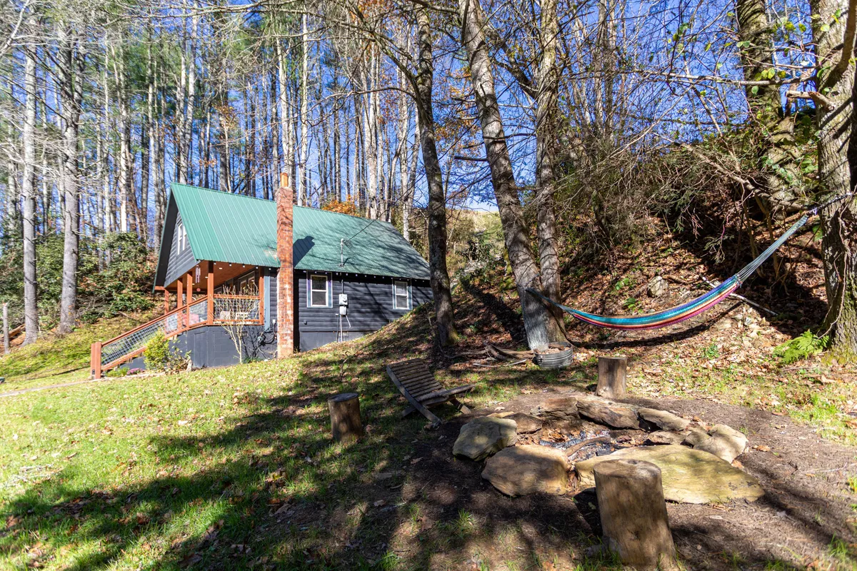 The image depicts the exterior of the Hidden Cabin nestled among tall trees, showcasing a welcoming hammock strung between two trees, a circular fire pit with benches, and the cozy porch of the cabin itself. Dappled sunlight filters through the woods, and the outdoor setting radiates tranquility while hinting at easy access to hiking or relaxing after a day of adventure.