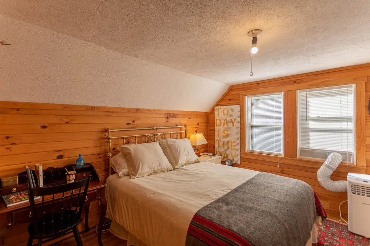This image showcases a beautiful queen bed against wooden walls, adorned with decorative pillows and tasteful wall art. The room is bathed in natural light streaming in from the windows, creating a serene and inviting atmosphere ideal for rest and relaxation. The elements suggest a master bedroom designed for privacy and comfort.