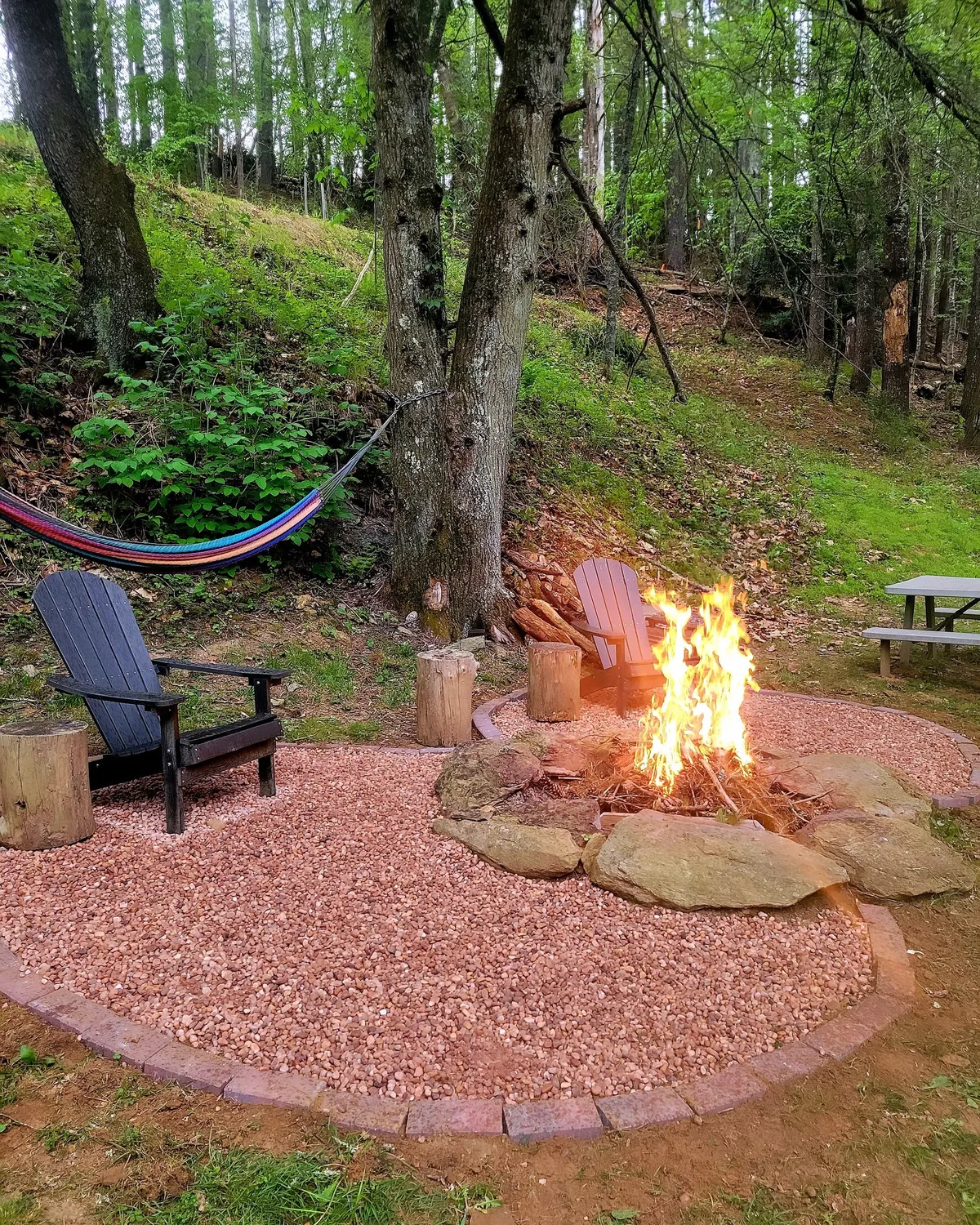 This image captures an inviting outdoor scene featuring a central fire pit surrounded by Adirondack chairs and a hammock nestled nearby. The natural setting evokes a sense of relaxation and togetherness, with ample space for people and pets to gather, play, and unwind after a day of exploring the mountains.