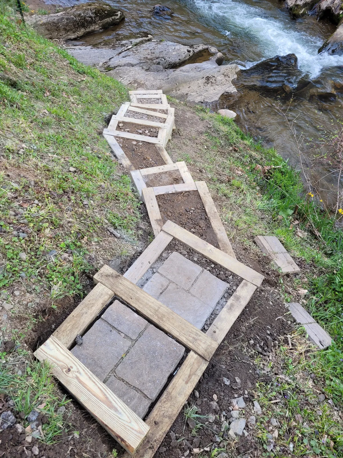 A wooden pathway meanders through lush greenery alongside a sparkling creek, evoking a sense of exploration and discovery. The surrounding plants and flowing water create an inviting scene for family walks, playful adventures, or quiet moments immersed in nature.