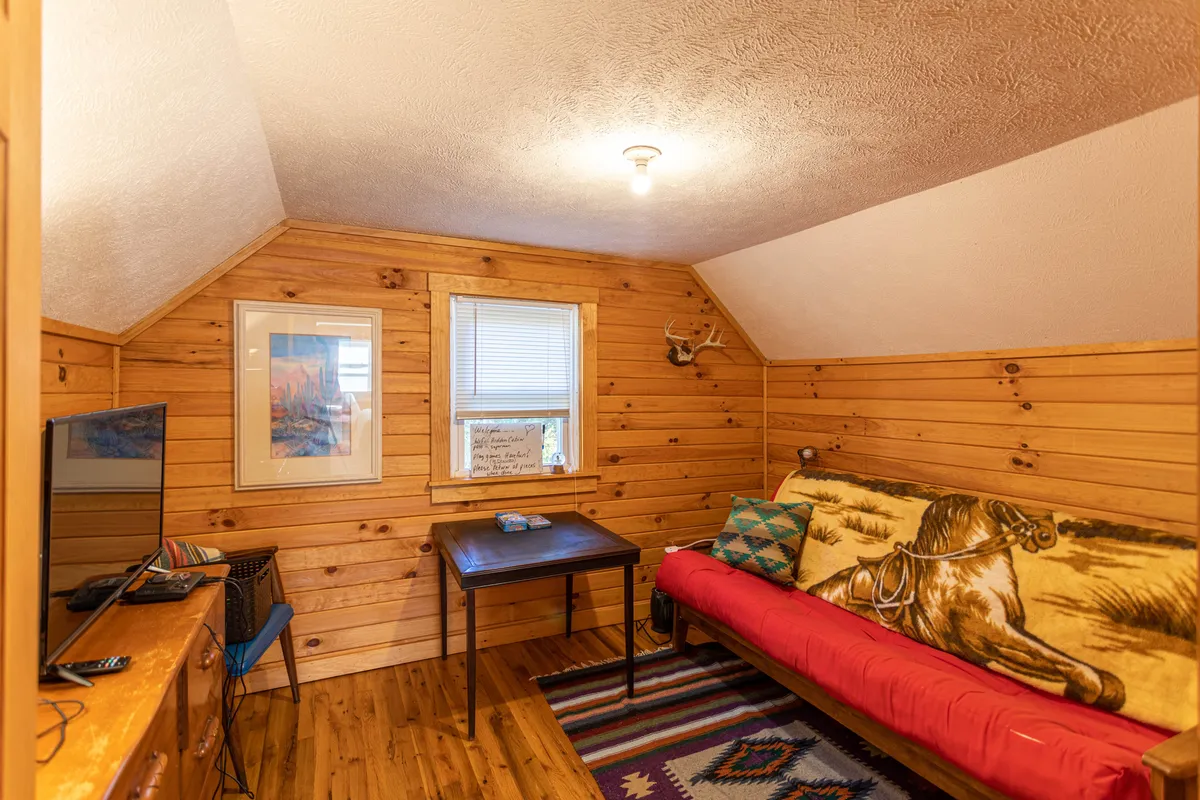 A cozy upstairs nook featuring a red couch draped with a horse blanket, wooden furniture, inviting artwork, and a textured rug underfoot. The arrangement suggests a relaxed, intimate atmosphere ideal for lounging, reading, or enjoying entertainment like TV or games.