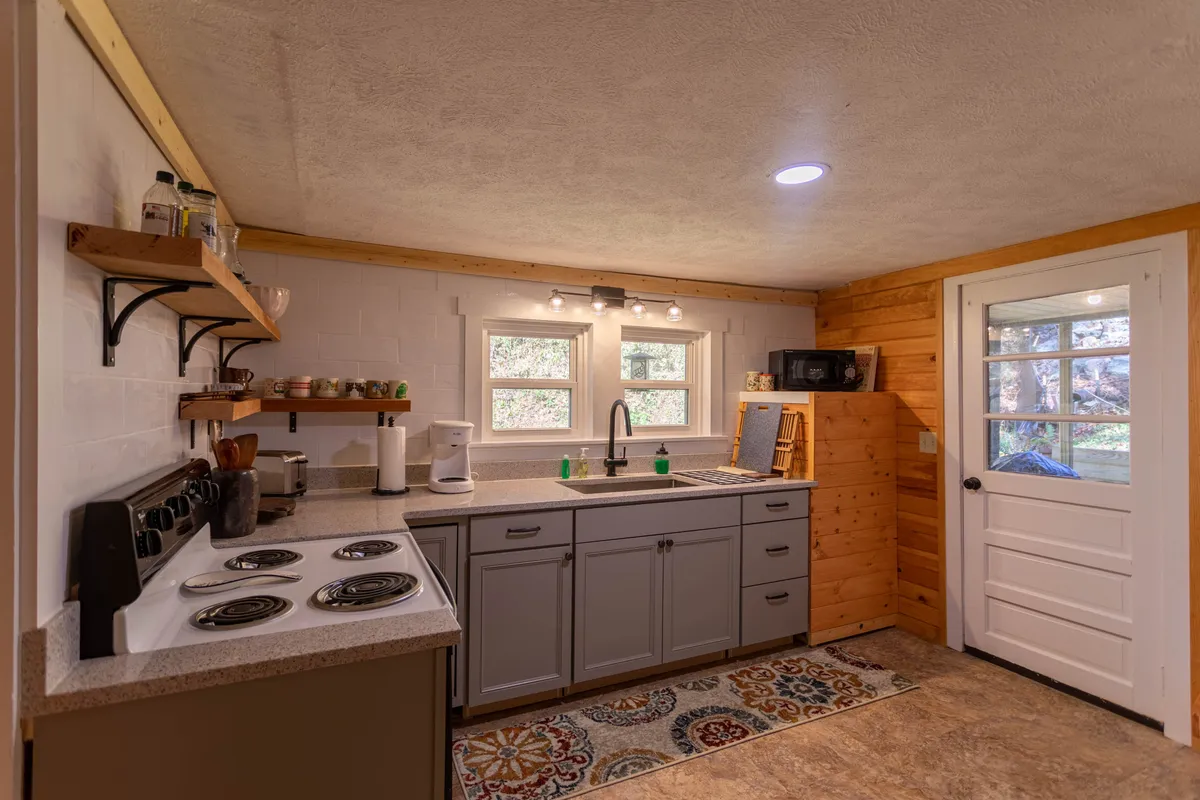 A warm, welcoming kitchen with a deep farmhouse sink, wooden shelves stocked with essentials, a sturdy cooking range, and sunlight streaming in through the window. The open layout hints at its connection to the dining and living areas, making it ideal for preparing meals while engaging with others.