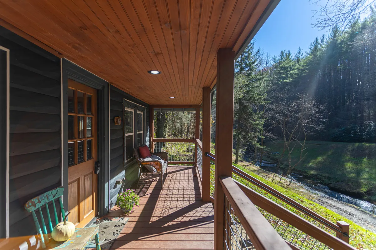 A wooden front porch with inviting rocking chairs overlooks a lush green yard and a gently flowing stream. The porch is partially shaded by trees, with hints of sunlight dappling the wooden boards. The tranquil water and greenery create a serene natural backdrop visible from the porch.