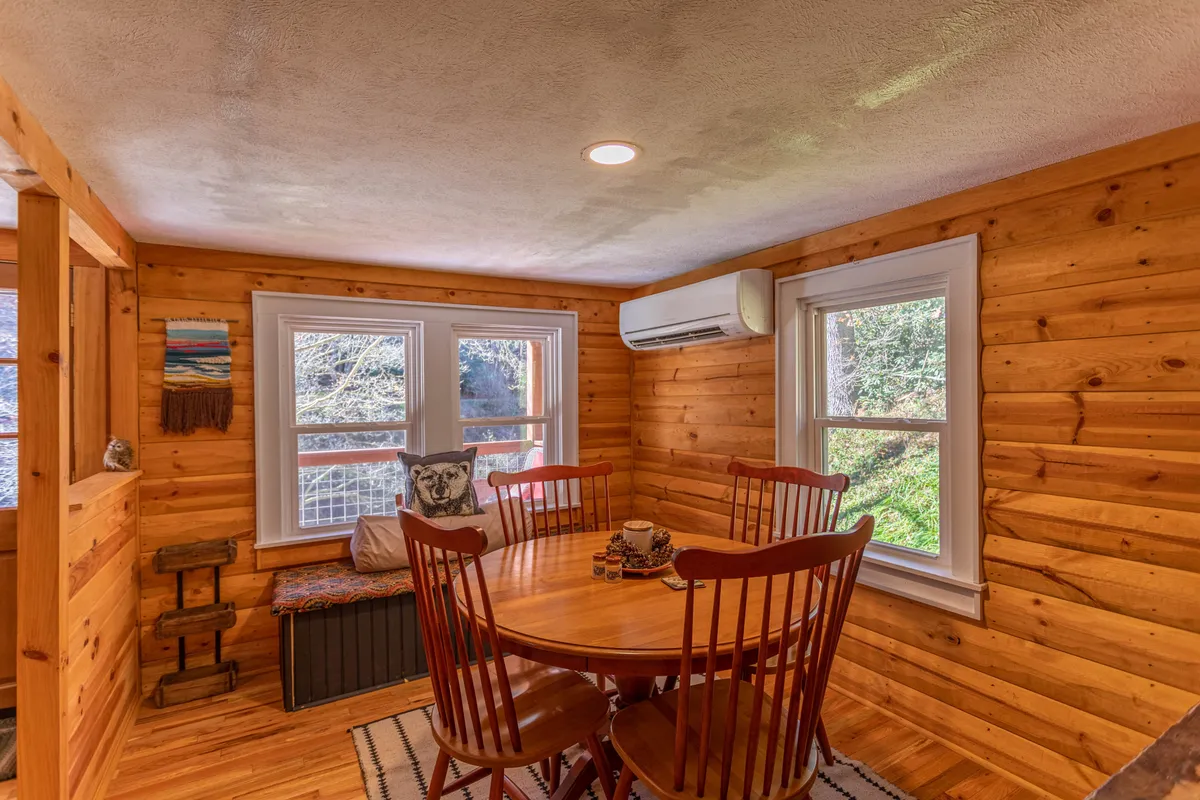 A cozy dining area featuring a round wooden table surrounded by matching chairs, set against warm wooden walls. Large windows invite natural light, and a bench seating nook adds to the sense of comfort and intimacy. The table is perfectly suited for meals, games, or leisurely conversations.